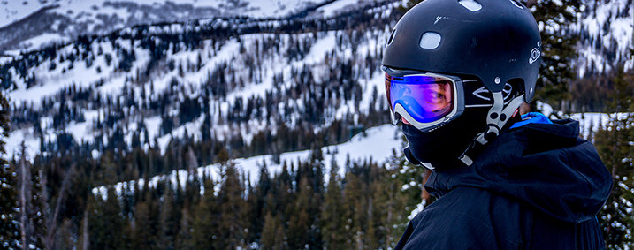 A skier in a ski mask and helmet standing in front of snow-covered pines as a backdrop